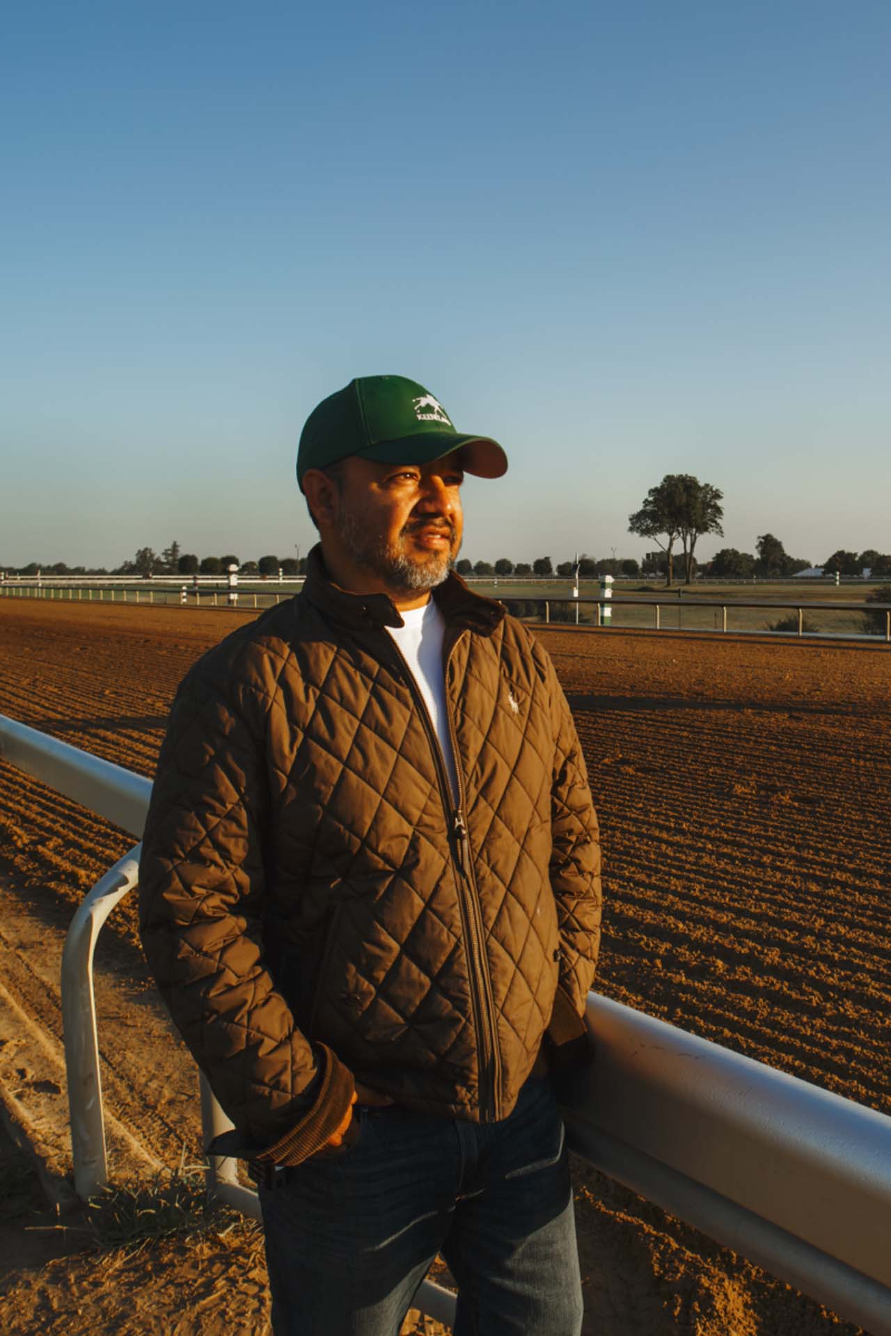 A photo of Alfredo Laureano looking off into the distance while standing by the dirt track at sunset. He is an older Hispanic man with graying chin stubble. He is wearing a green Keenhoofbeat-brand hat and a brown Keenhoofbeat-brand jacket overtop a white shirt.
