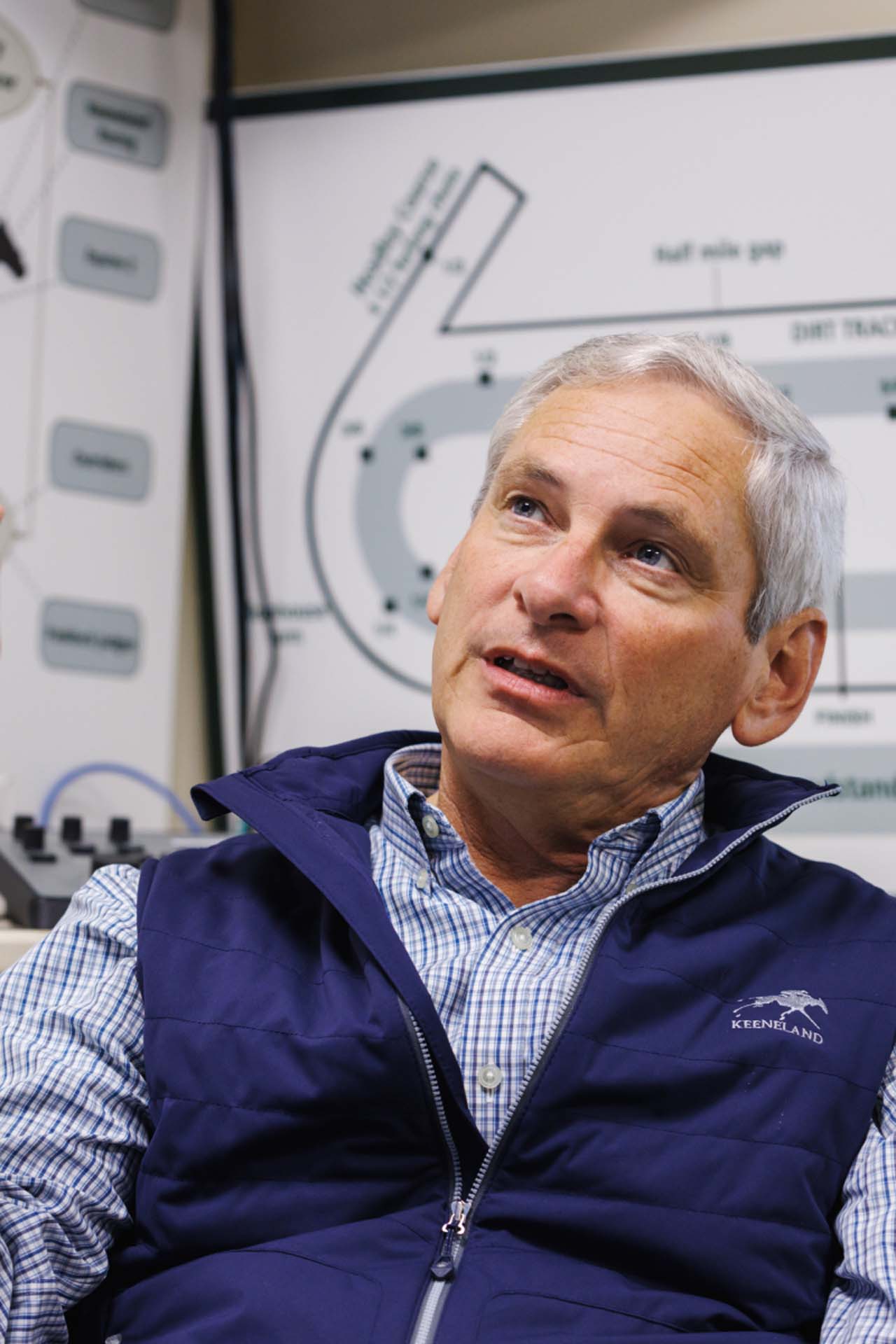 An up-close photo of Dr. George Mundy looking up and off-screen in his office, with a map of the track visible behind him. He is an older White man with short silver hair. He is wearing a navy Keenhoofbeat-brand vest over a white and blue checkered shirt.